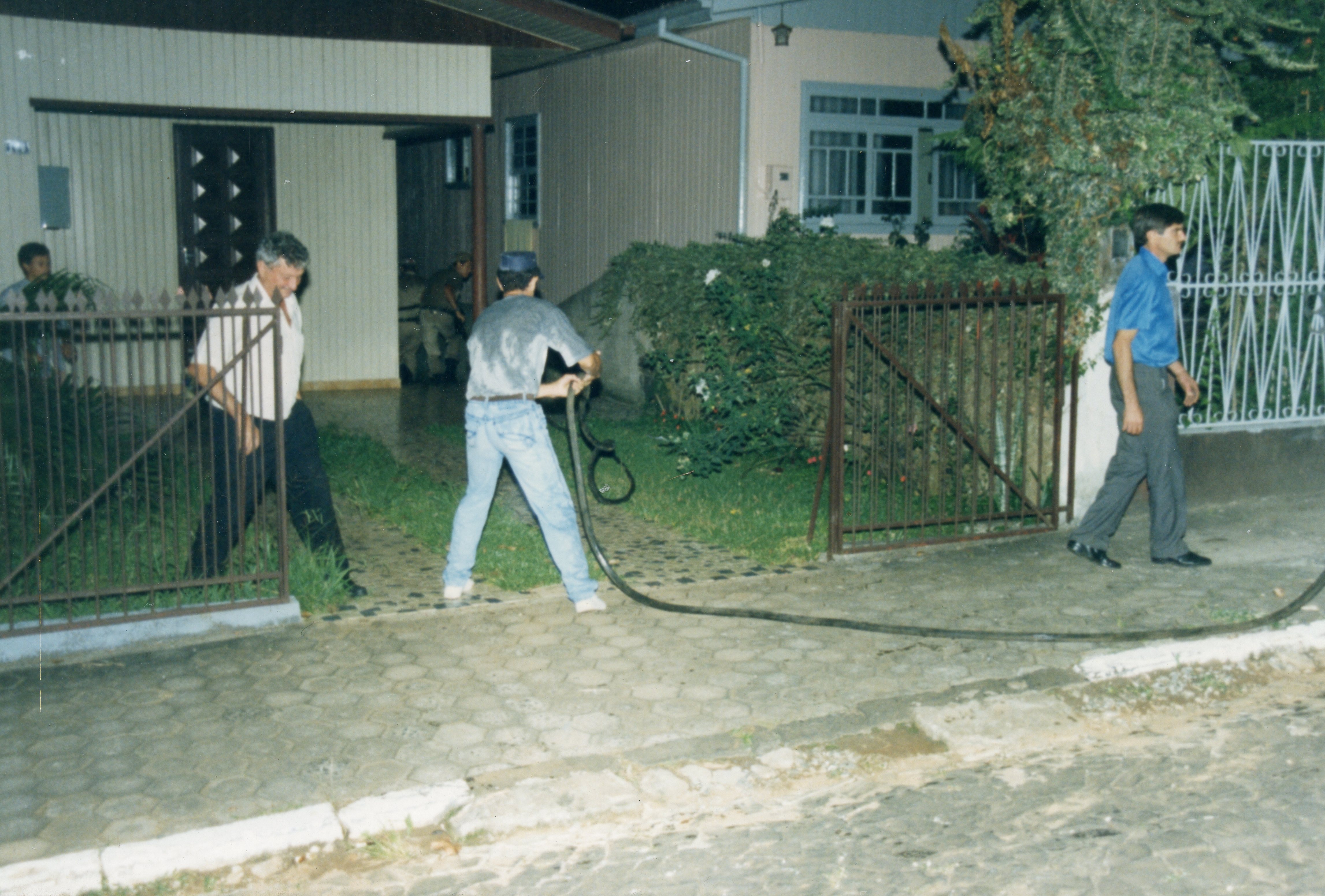 Uma cena cotidiana capturada em frente a uma residência, onde homens manuseiam uma mangueira, possivelmente realizando uma tarefa de limpeza ou manutenção. A vestimenta e a arquitetura das casas sugerem um cenário brasileiro típico das décadas de 1980 ou 1990.