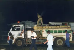 Uma fotografia noturna que registra um caminhão de serviço, possivelmente de bombeiros ou manutenção, com um homem operando equipamentos no topo do veículo. A cena apresenta pessoas caminhando ao redor do caminhão em um ambiente urbano, sugerindo uma atividade de serviço público ou emergência em uma cidade brasileira, provavelmente entre as décadas de 1980 e 1990.