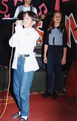 Uma fotografia de um evento musical, possivelmente um show escolar ou festival local, apresentando jovens cantores no palco. O estilo das roupas, como o colete xadrez e o corte de cabelo, sugere fortemente a estética da década de 1990.