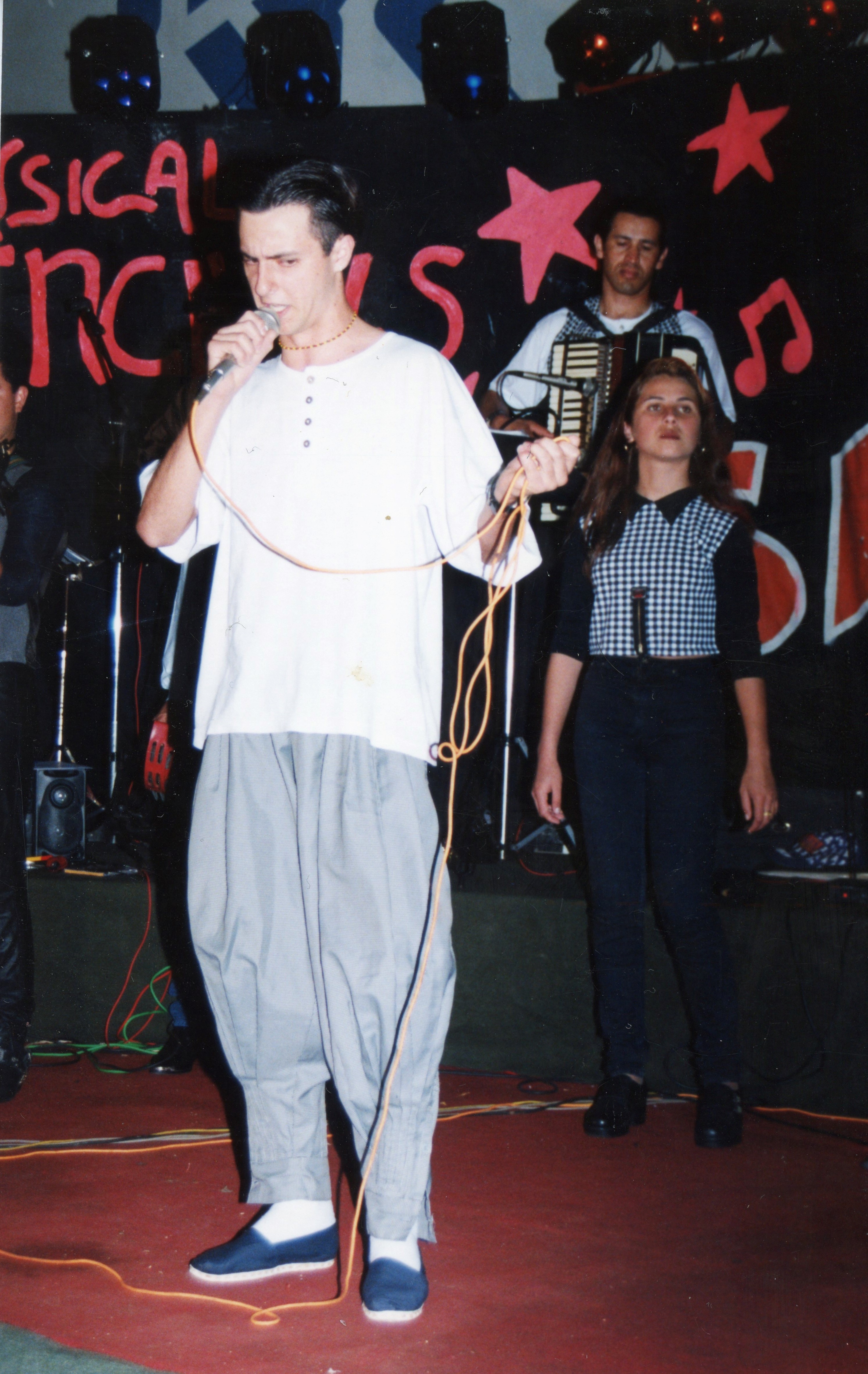 Uma fotografia de uma banda musical em um palco, com um vocalista em primeiro plano segurando um microfone, acompanhado por uma mulher e um sanfoneiro ao fundo. O cenário apresenta uma decoração com estrelas e letras pintadas, sugerindo um evento musical ou festival local típico da década de 1990.