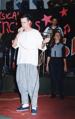 Uma fotografia de uma banda musical em um palco, com um vocalista em primeiro plano segurando um microfone, acompanhado por uma mulher e um sanfoneiro ao fundo. O cenário apresenta uma decoração com estrelas e letras pintadas, sugerindo um evento musical ou festival local típico da década de 1990.