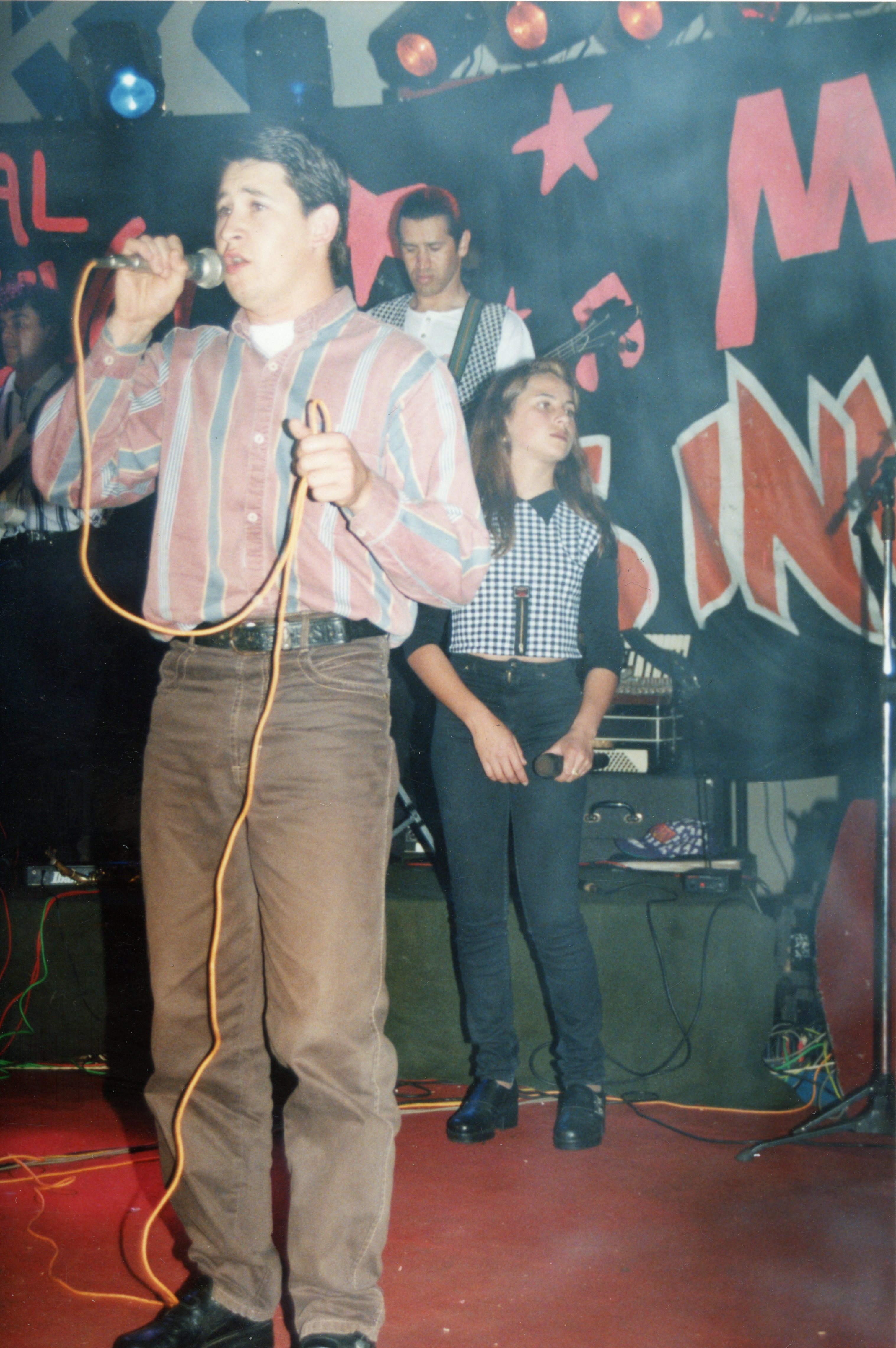 Uma fotografia de uma banda musical em um palco, provavelmente em um evento local ou festa popular brasileira. Os jovens vestem roupas típicas da década de 1990, com camisas listradas e calças jeans, enquanto cantam e tocam instrumentos diante de um painel decorativo com estrelas e letras pintadas.