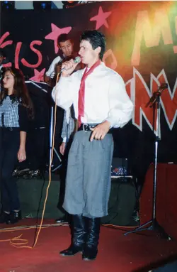 Uma fotografia colorida capturando uma apresentação musical em um palco, possivelmente um evento regional ou festival de música tradicionalista. O cantor principal veste uma camisa branca com lenço vermelho e bombachas, elementos típicos da indumentária gaúcha, sugerindo um contexto cultural do sul do Brasil na década de 1990.