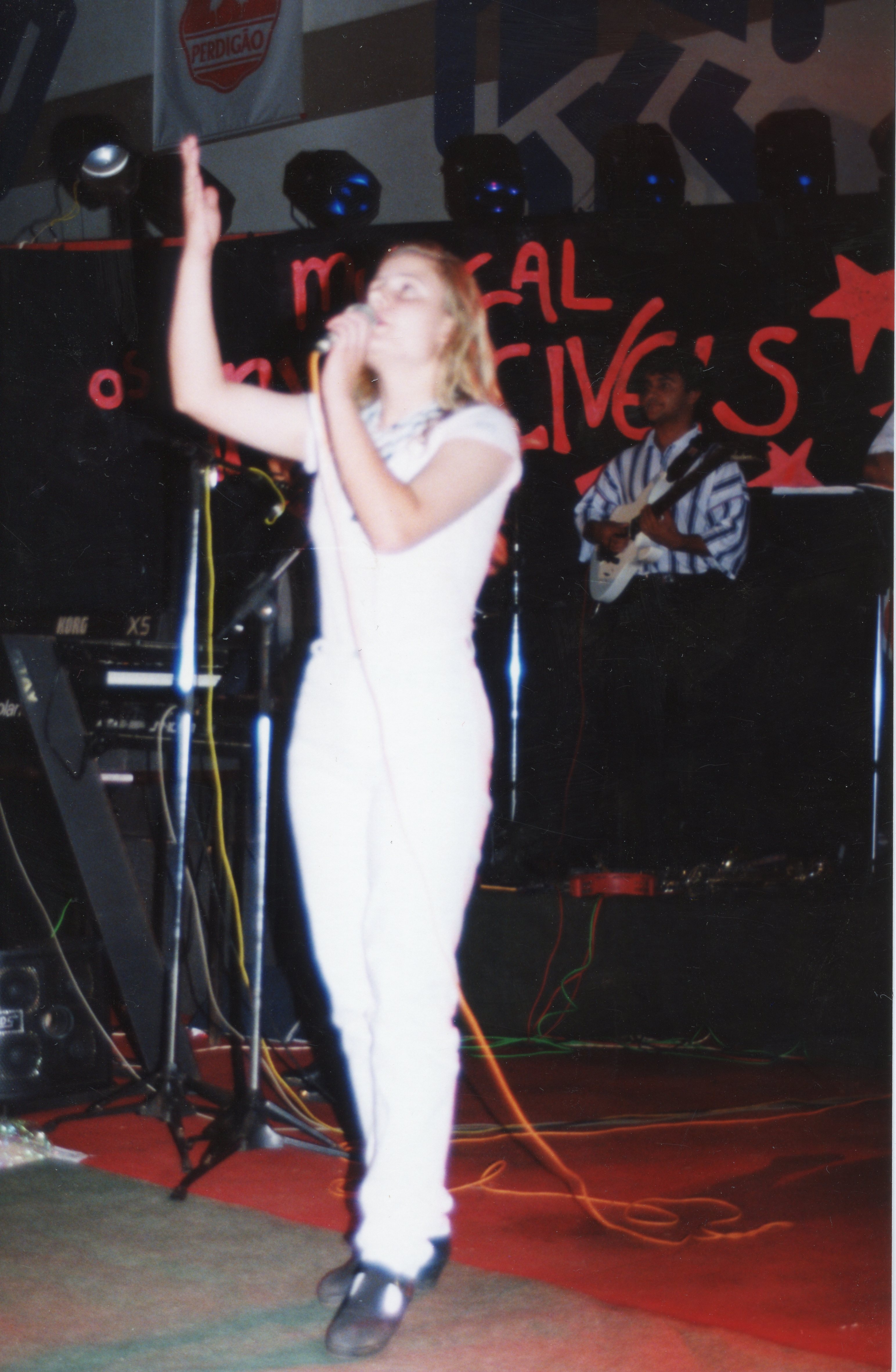 Uma fotografia de uma apresentação musical ao vivo, possivelmente em um evento corporativo ou comunitário, com uma cantora em destaque no palco. Ao fundo, vê-se um músico tocando guitarra e um banner com a marca 'Perdigão', sugerindo um contexto de evento em uma cidade com forte presença industrial, como Videira.