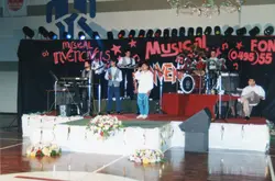 Uma banda musical chamada 'Os Invencíveis' se apresenta em um palco decorado com arranjos de flores e um painel de fundo com o nome do grupo. A cena, provavelmente datada da década de 1990, mostra músicos com instrumentos como teclado, bateria e microfones em um ambiente que parece ser um ginásio ou salão de eventos.