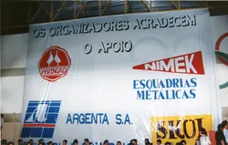 Uma faixa de grandes dimensões exibida em um ambiente interno, provavelmente um ginásio, contendo logotipos de empresas locais e regionais como Perdigão, Nimek, Argenta S.A. e Skol. O banner expressa agradecimento aos patrocinadores e apoiadores de um evento, refletindo a cultura corporativa e o apoio empresarial a atividades comunitárias típicas de cidades como Videira.