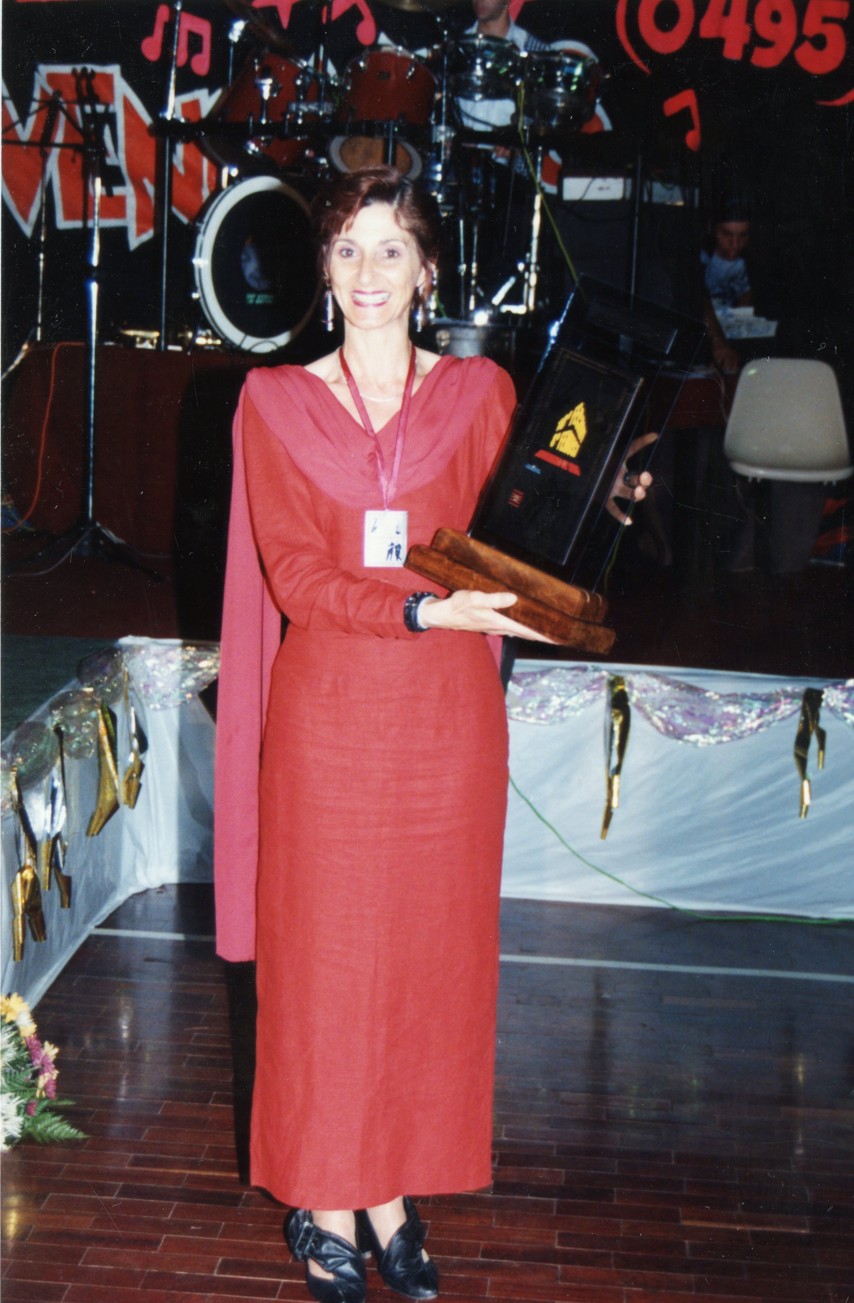 Uma mulher sorridente, vestindo um longo vestido vermelho, segura um troféu em um palco com instrumentos musicais ao fundo. A cena sugere uma cerimônia de premiação ou evento social, possivelmente ocorrido entre o final da década de 1990 e início dos anos 2000, dado o estilo da fotografia e do vestuário.