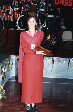 Uma mulher sorridente, vestindo um longo vestido vermelho, segura um troféu em um palco com instrumentos musicais ao fundo. A cena sugere uma cerimônia de premiação ou evento social, possivelmente ocorrido entre o final da década de 1990 e início dos anos 2000, dado o estilo da fotografia e do vestuário.