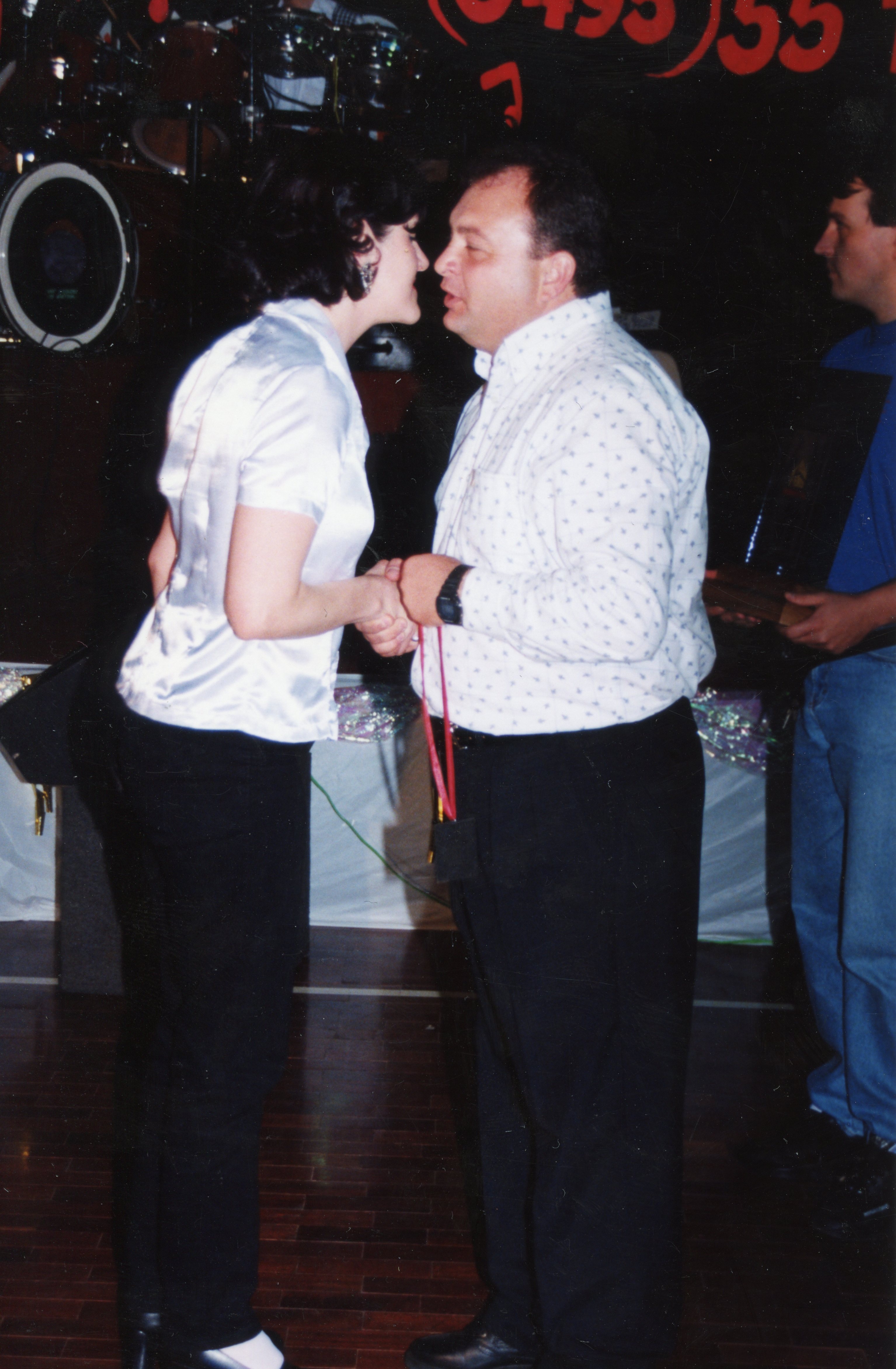 Uma fotografia que captura um momento de interação social, possivelmente uma premiação ou cumprimento formal, entre um homem e uma mulher em um ambiente interno. O cenário sugere um evento noturno, com equipamentos de som ao fundo, típico de festividades ou celebrações comunitárias, com vestimentas que remetem ao final da década de 1990 ou início dos anos 2000.