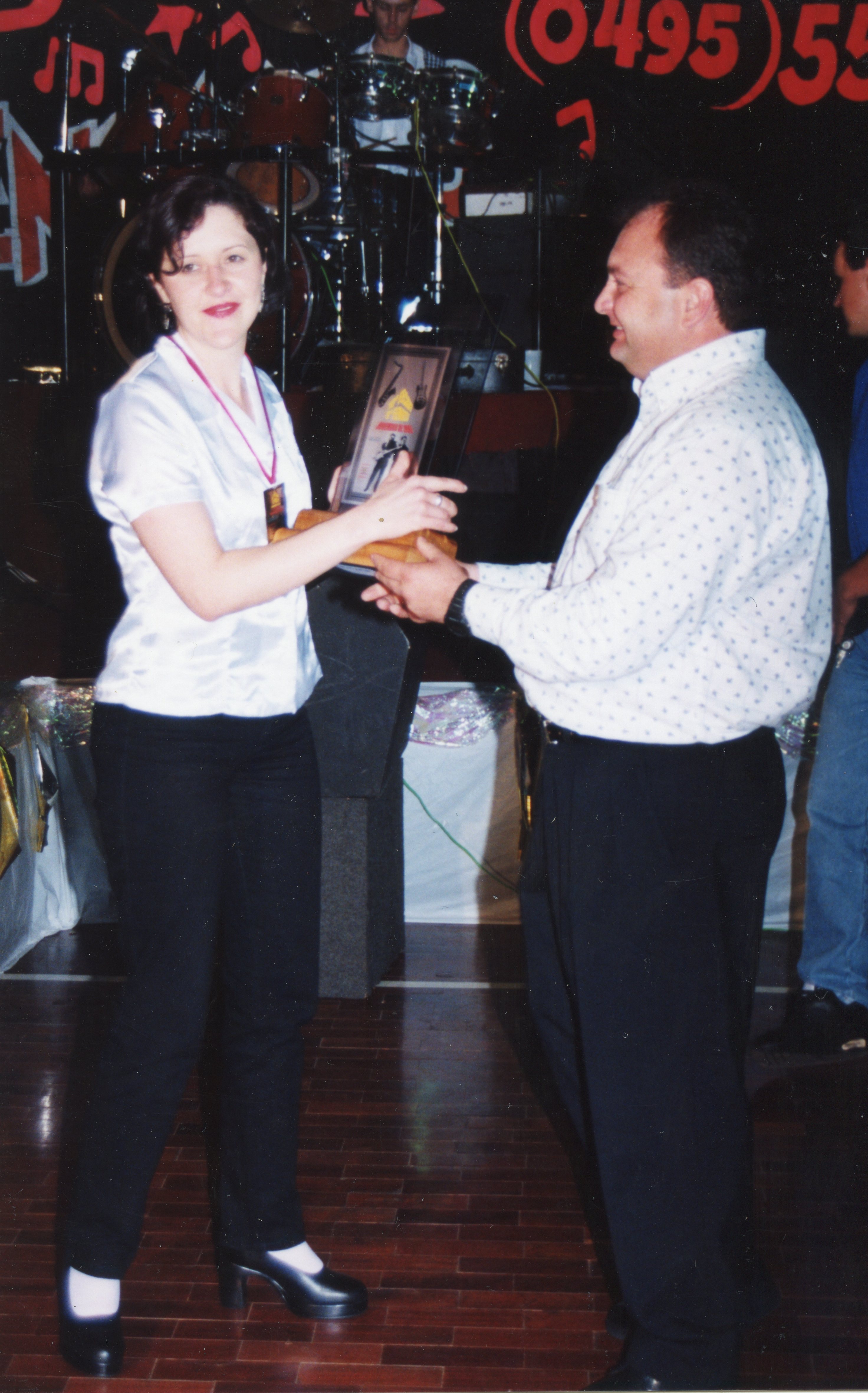 Uma fotografia de um evento social onde uma mulher entrega um troféu ou placa comemorativa a um homem. O cenário sugere um ambiente de palco ou salão de festas, com instrumentos musicais ao fundo e vestimentas típicas do final da década de 1990 ou início dos anos 2000.