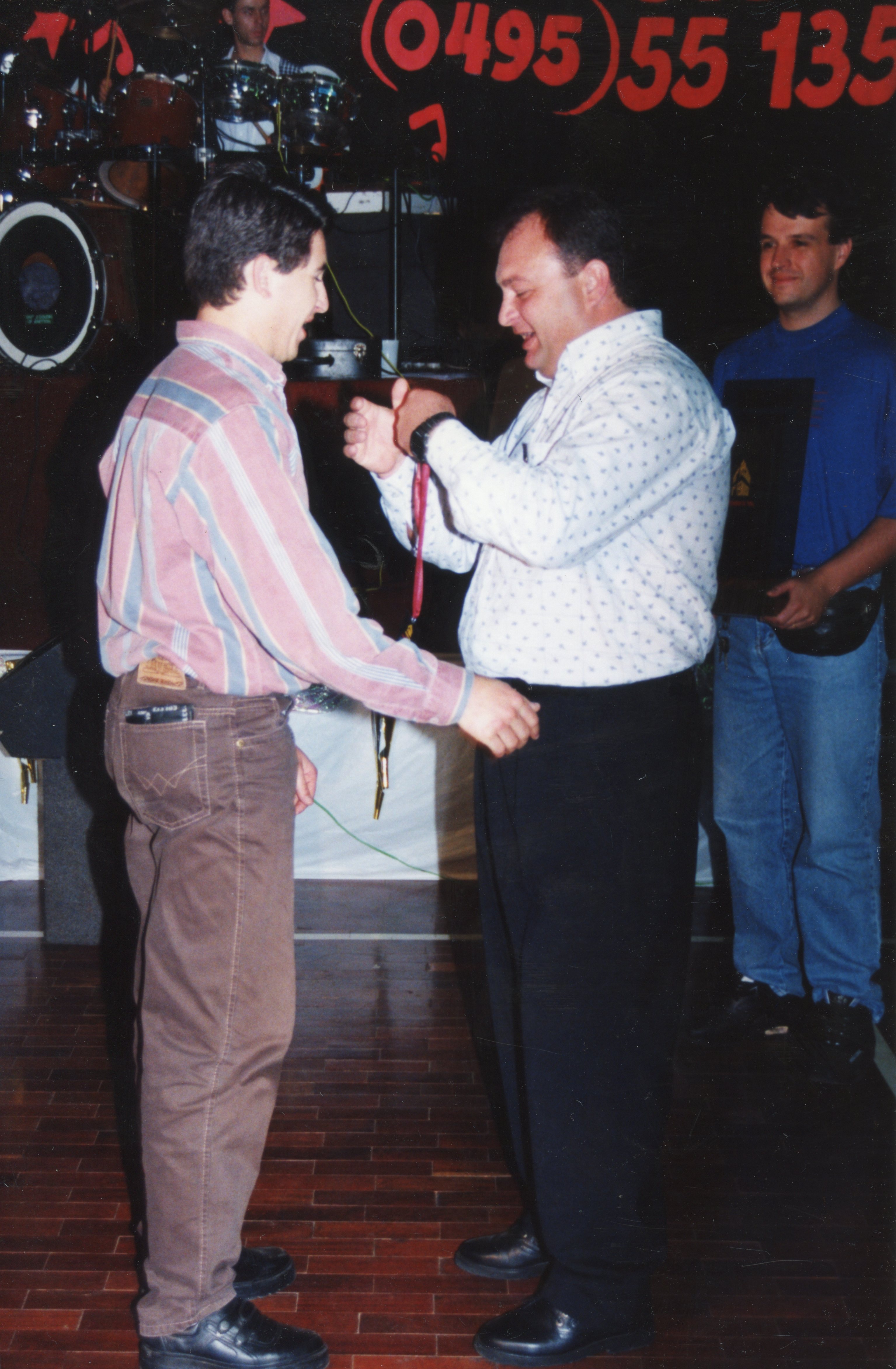 Uma fotografia de um evento social ou cerimônia de premiação em ambiente interno, onde um homem entrega uma medalha ou crachá a outro. O estilo das vestimentas, como camisas de botões e calças jeans, sugere uma estética típica da década de 1990, com um código de área visível ao fundo indicando uma possível origem brasileira.