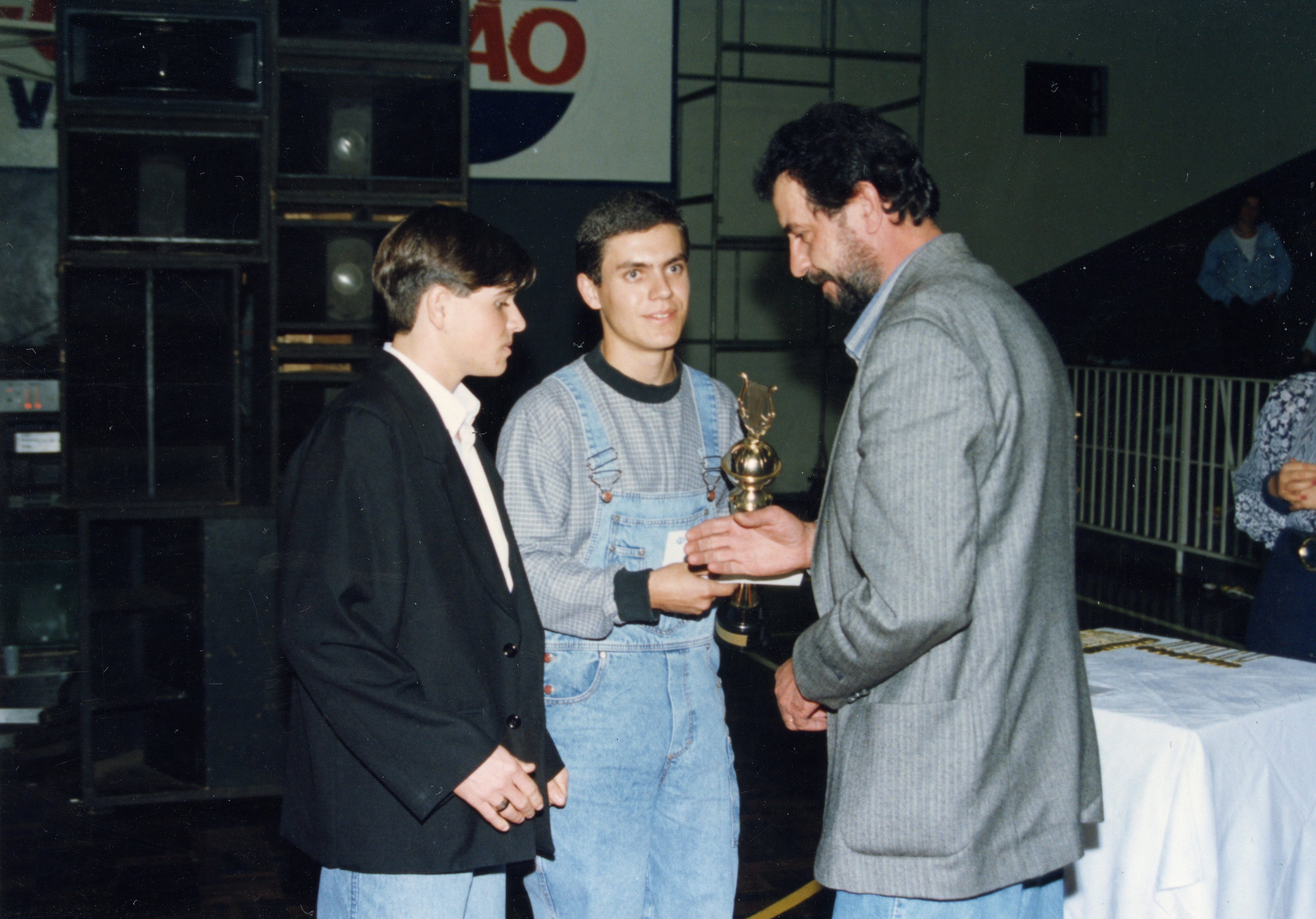 Uma fotografia colorida que registra uma cerimônia de premiação, onde um homem adulto entrega um troféu a um jovem que veste um macacão jeans. A cena ocorre em um ambiente interno, possivelmente um ginásio ou salão de eventos, com equipamentos de som ao fundo, sugerindo um evento esportivo ou cultural da década de 1990.