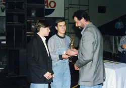 Uma fotografia colorida que registra uma cerimônia de premiação, onde um homem adulto entrega um troféu a um jovem que veste um macacão jeans. A cena ocorre em um ambiente interno, possivelmente um ginásio ou salão de eventos, com equipamentos de som ao fundo, sugerindo um evento esportivo ou cultural da década de 1990.