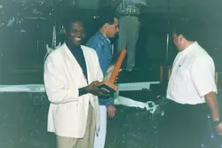 Uma fotografia que registra um momento de premiação ou homenagem, onde um homem sorridente recebe uma placa de madeira. O cenário sugere um evento social ou corporativo em um ambiente fechado, com vestimentas típicas das décadas de 1990 ou início de 2000.