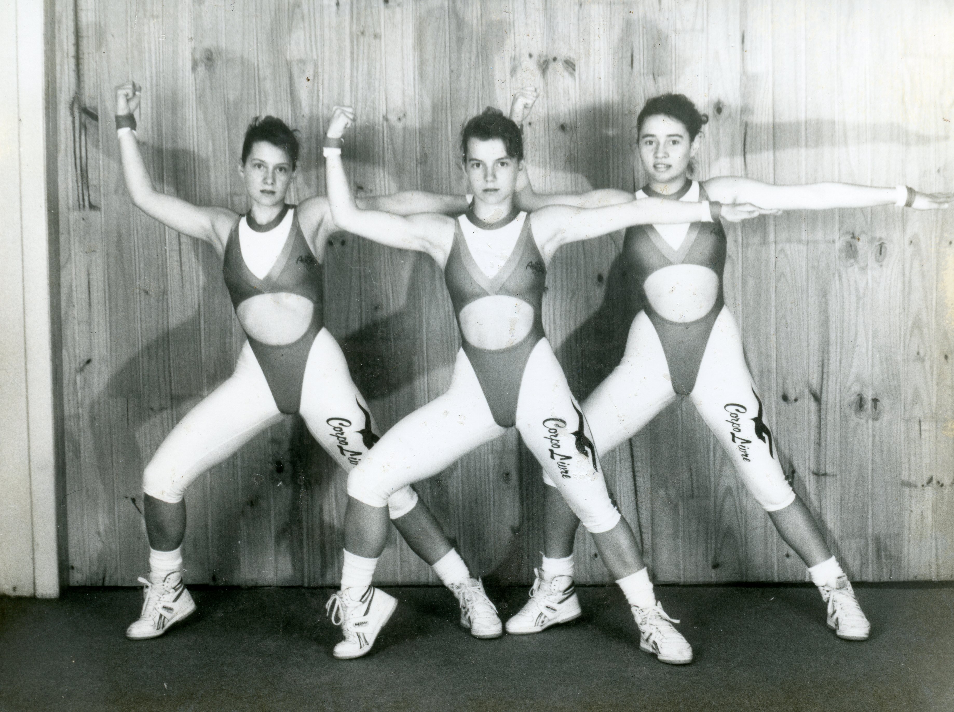 Fotografia em preto e branco de três jovens mulheres posando em uma coreografia de ginástica ou aeróbica, vestindo collants esportivos com a inscrição 'Corpo Livre'. A imagem reflete a estética fitness popular no final do século XX, possivelmente em um ambiente de academia ou estúdio de dança.