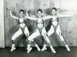 Fotografia em preto e branco de três jovens mulheres posando em uma coreografia de ginástica ou aeróbica, vestindo collants esportivos com a inscrição 'Corpo Livre'. A imagem reflete a estética fitness popular no final do século XX, possivelmente em um ambiente de academia ou estúdio de dança.