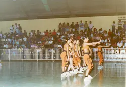 Fotografia de um grupo de ginastas ou dançarinas vestindo uniformes esportivos laranja e preto, realizando uma apresentação em uma quadra coberta. O evento conta com a presença de um público numeroso nas arquibancadas, sugerindo uma atividade esportiva ou cultural comunitária, possivelmente na década de 1990.