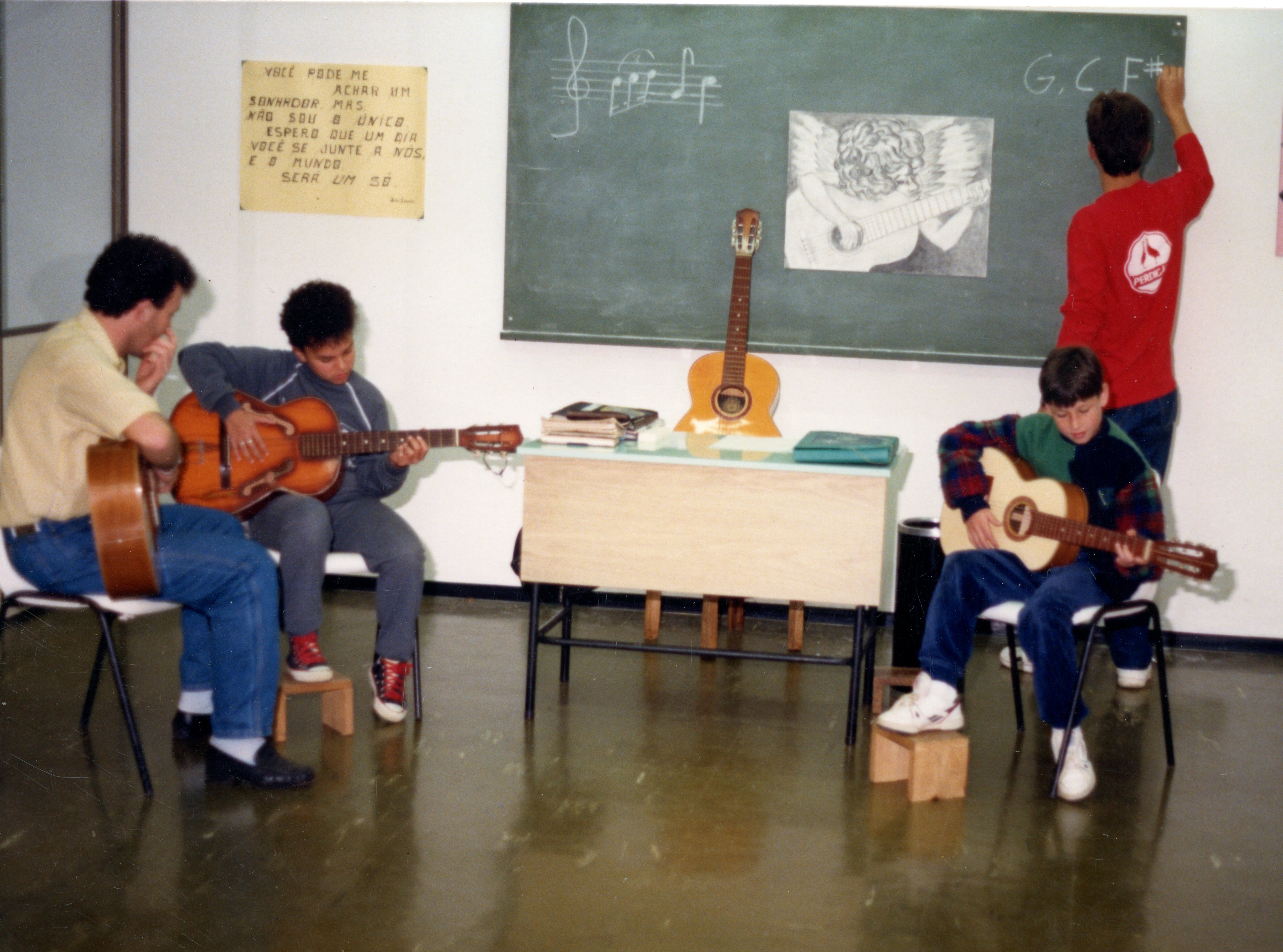 Uma cena de sala de aula de música onde três jovens tocam violão enquanto um quarto aluno escreve notas musicais no quadro-negro. O ambiente apresenta elementos típicos de uma escola ou centro cultural, com um cartaz com a letra de 'Imagine' de John Lennon na parede, sugerindo um contexto educacional ou de lazer da década de 1990.