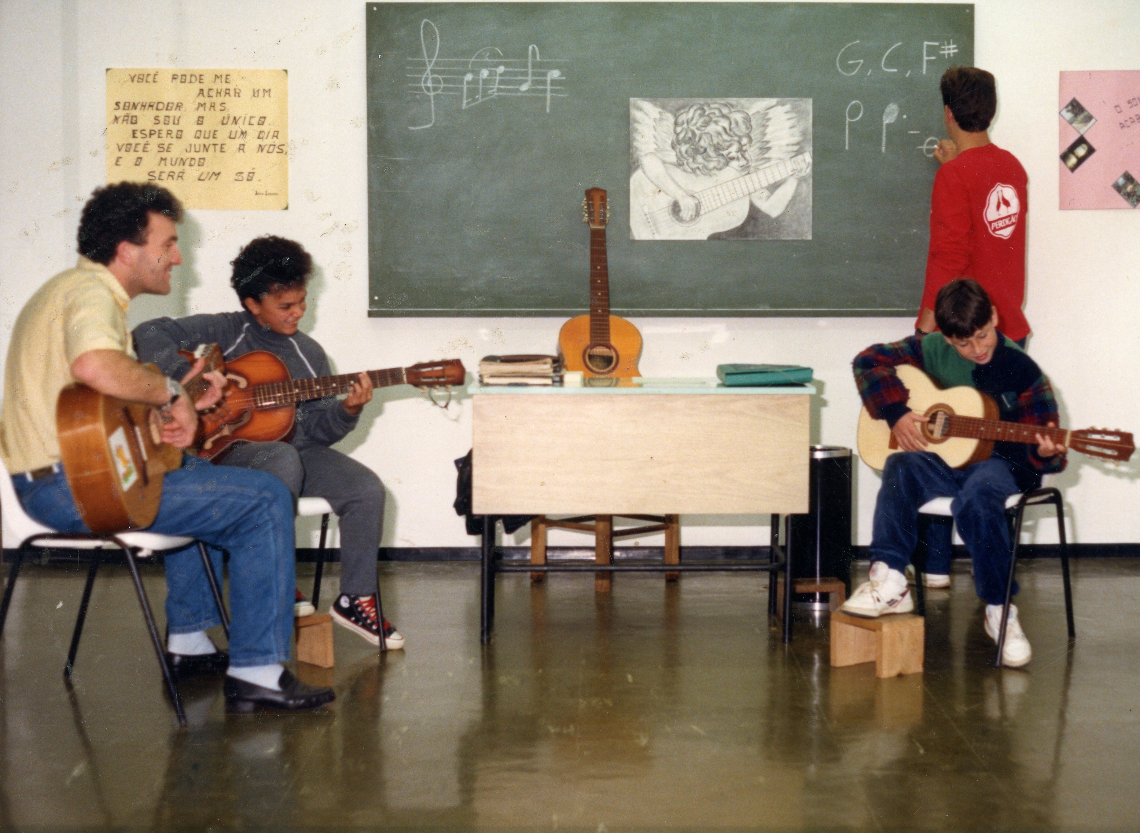 Uma cena de uma aula de música em uma sala de aula, provavelmente na década de 1990, com três pessoas tocando violão e um aluno escrevendo na lousa. O ambiente apresenta elementos típicos de escola, como lousa com partituras e desenhos, e cartazes com mensagens inspiradoras, sugerindo uma atividade cultural ou educacional.