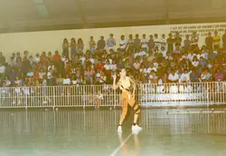 Uma fotografia colorida capturando uma apresentação de ginástica ou dança em uma quadra esportiva coberta, possivelmente em um ginásio municipal. Uma jovem atleta em destaque veste um collant esportivo, enquanto uma grande plateia assiste das arquibancadas ao fundo, sob uma inscrição pintada na parede.