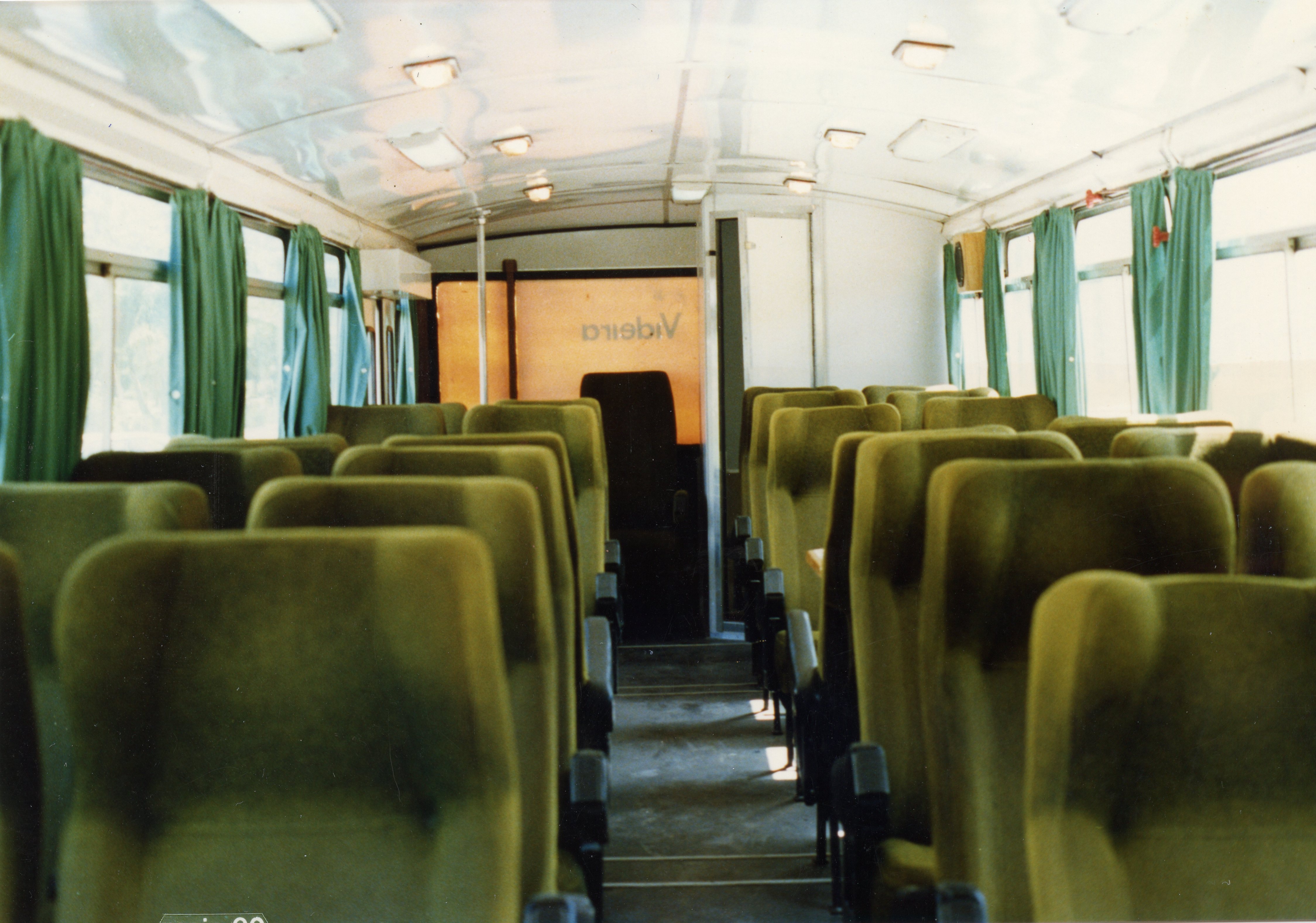 Interior de um ônibus de viagem antigo, apresentando poltronas estofadas em tom esverdeado e cortinas da mesma cor. Ao fundo, é possível ler a palavra 'Videira' em uma divisória, sugerindo uma conexão com a cidade catarinense, possivelmente em um veículo de transporte intermunicipal das décadas de 1970 ou 1980.