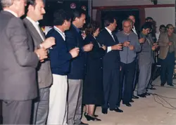 Uma fotografia de um grupo de pessoas em um evento social ou corporativo, possivelmente uma confraternização, onde os participantes seguram taças. O estilo das vestimentas, incluindo ternos e blazers, sugere um contexto brasileiro das décadas de 1980 ou 1990, com a presença de logotipos da marca Perdigão ao fundo.