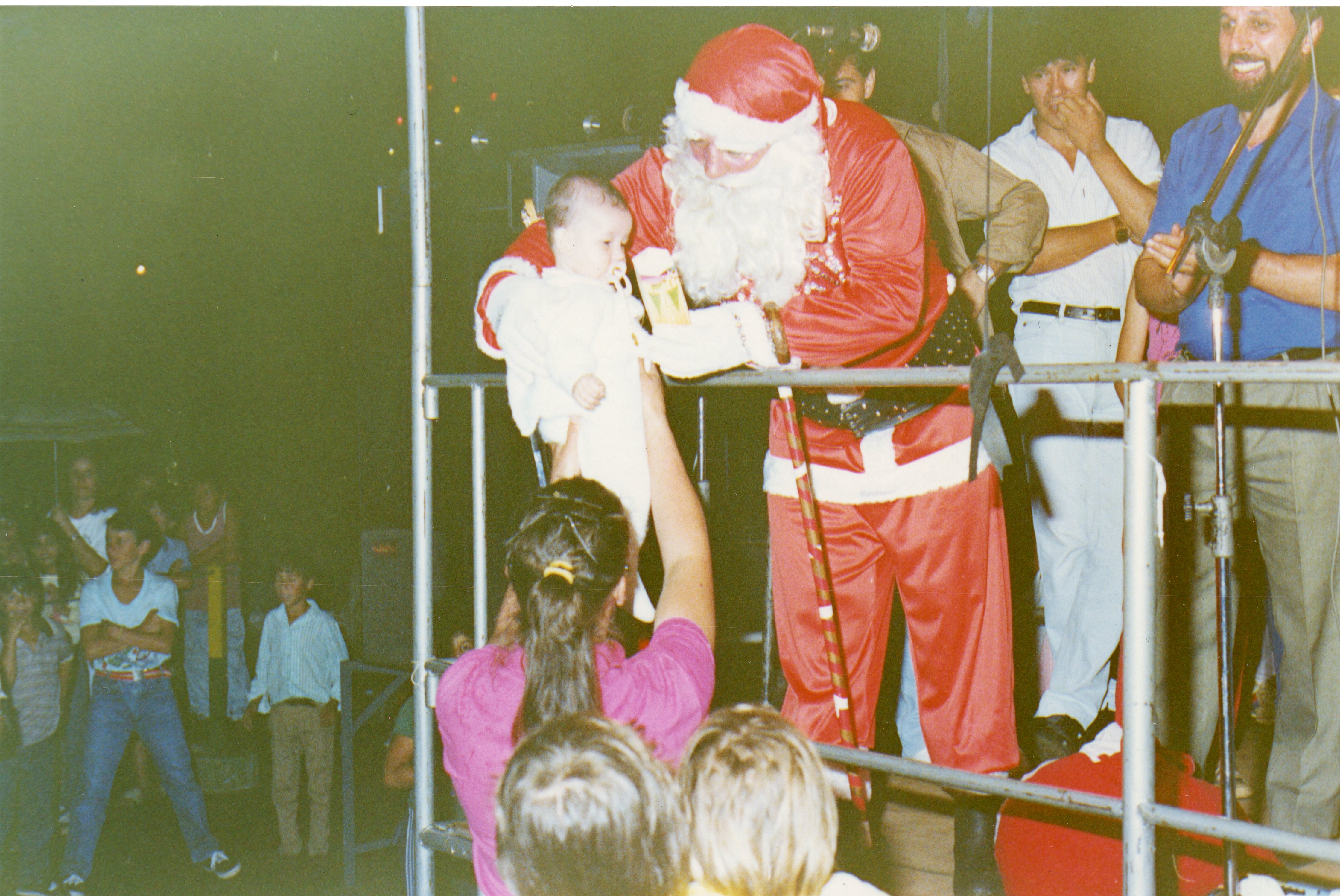 Uma fotografia colorida que captura um evento comunitário de Natal, onde uma pessoa fantasiada de Papai Noel interage com um bebê em um palco improvisado. O ambiente sugere uma celebração pública noturna em uma pequena cidade brasileira, com espectadores observando ao fundo e uma estrutura metálica de palco visível.