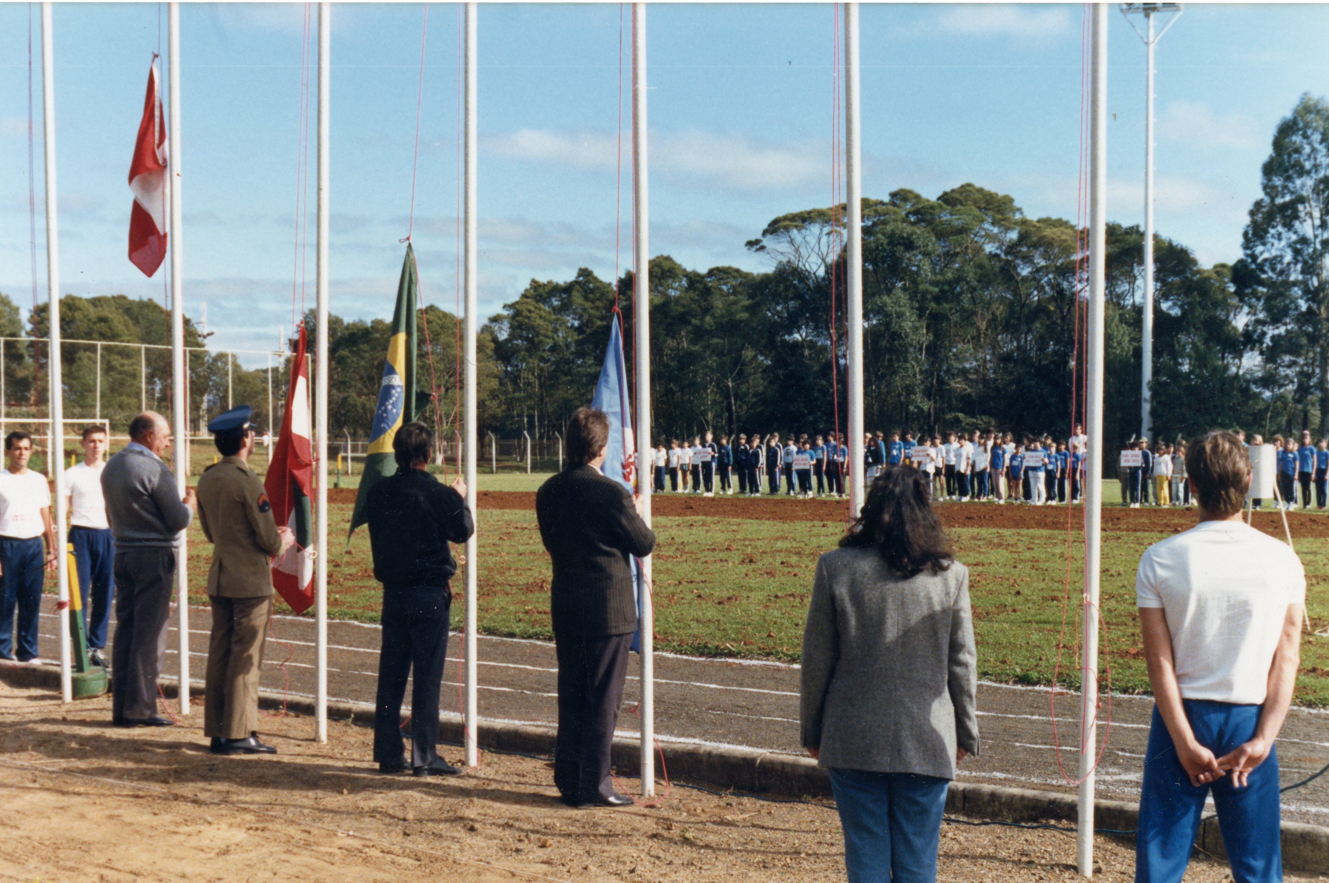 Uma cerimônia cívica ou esportiva ao ar livre em um campo com pista de atletismo, onde várias pessoas estão perfiladas ao fundo. Em primeiro plano, autoridades e participantes observam o hasteamento de bandeiras, incluindo a bandeira do Brasil, sugerindo um evento oficial ou escolar.