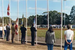 Uma cerimônia cívica ou esportiva ao ar livre em um campo com pista de atletismo, onde várias pessoas estão perfiladas ao fundo. Em primeiro plano, autoridades e participantes observam o hasteamento de bandeiras, incluindo a bandeira do Brasil, sugerindo um evento oficial ou escolar.