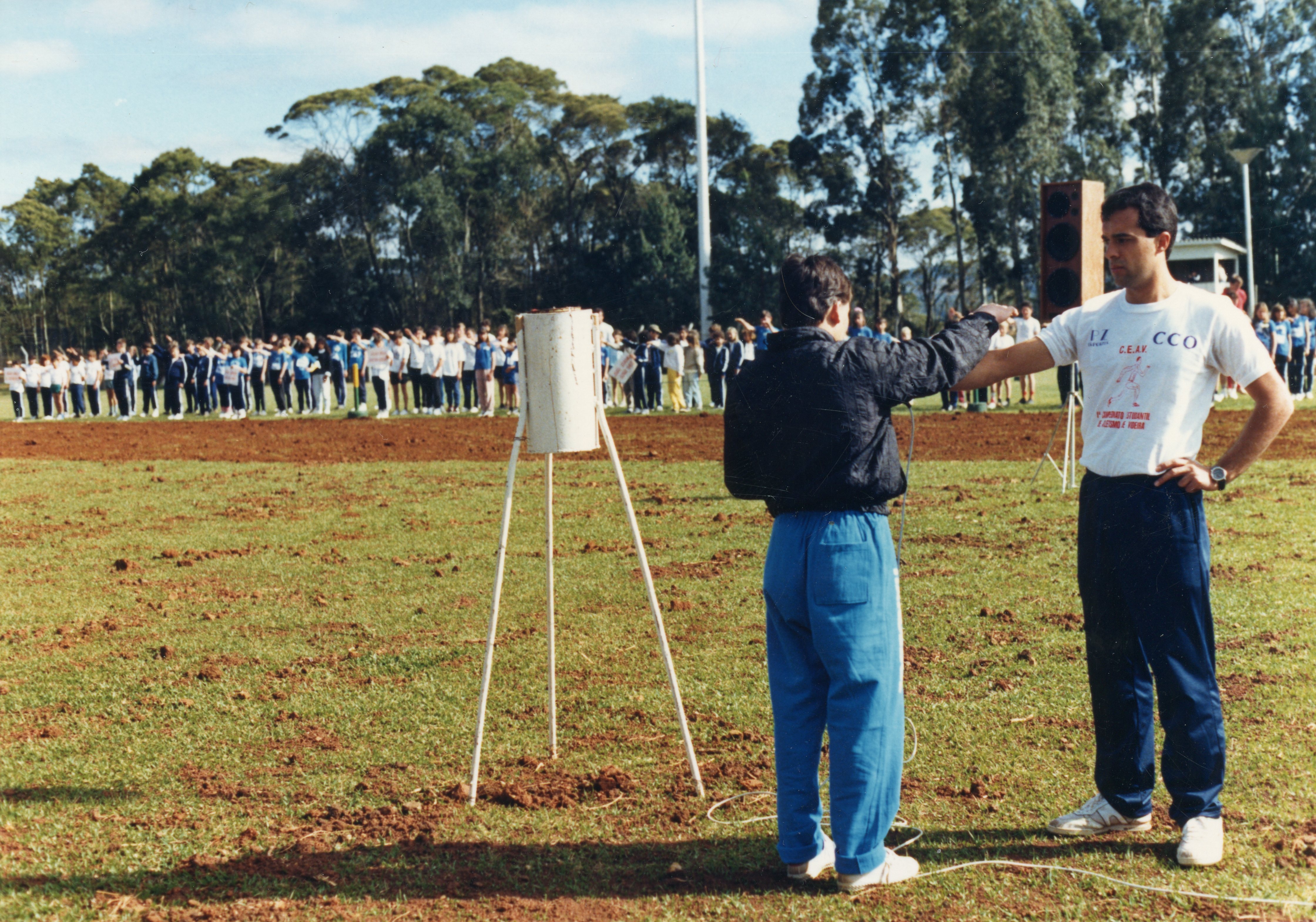 Uma fotografia que registra um evento esportivo ou cívico ao ar livre, possivelmente um desfile escolar ou competição atlética em um campo de terra batida. Em primeiro plano, dois homens organizam o evento, enquanto ao fundo uma longa fila de estudantes perfilados observa a cerimônia, sugerindo um contexto de organização escolar ou municipal típica de cidades do interior brasileiro.