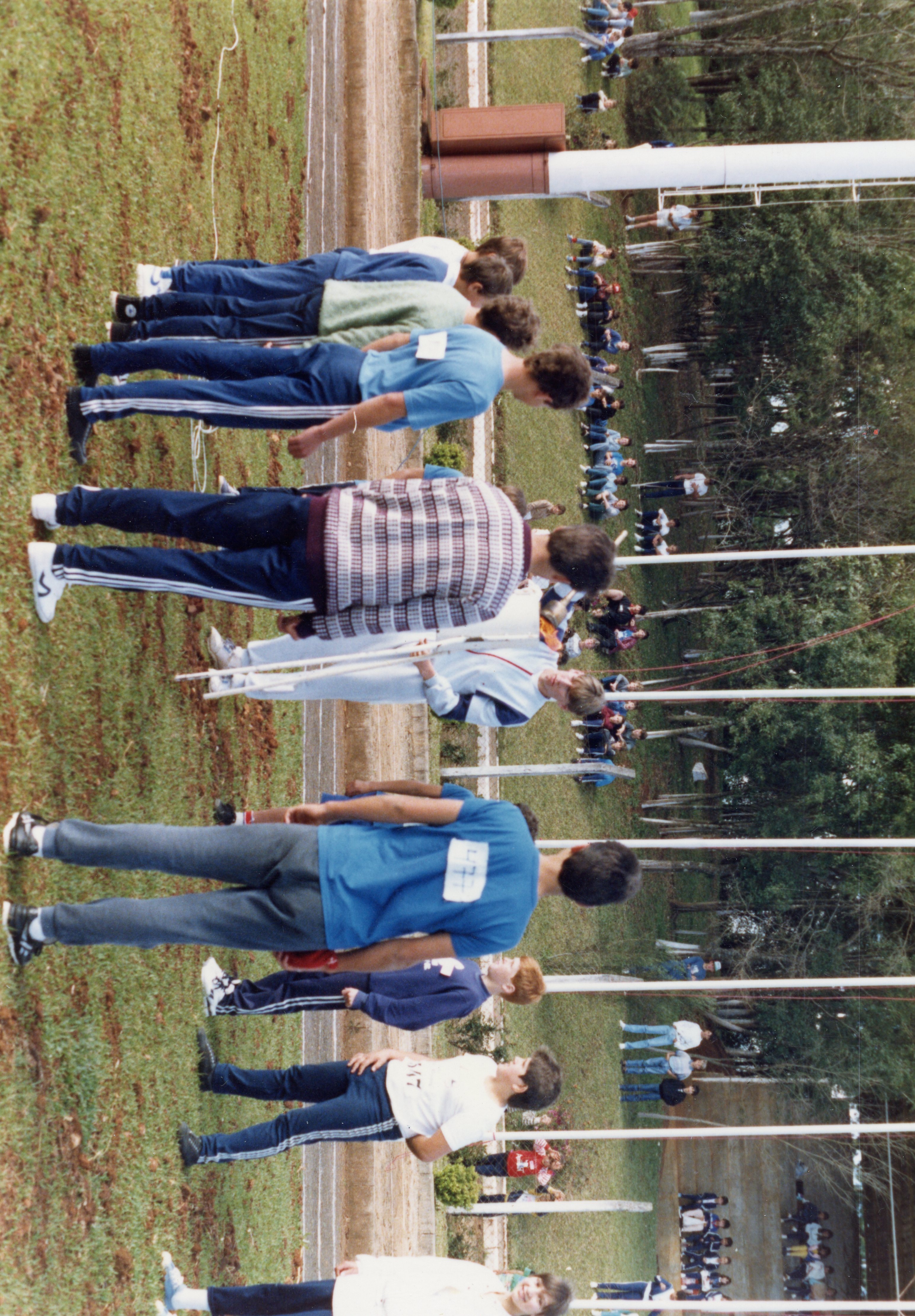 Uma fotografia de um evento esportivo escolar ao ar livre, possivelmente uma competição de atletismo, com diversos jovens alinhados em uma pista de terra. Os participantes vestem roupas esportivas típicas do final do século XX, como agasalhos e camisetas com números de identificação, em um ambiente rural ou de parque.