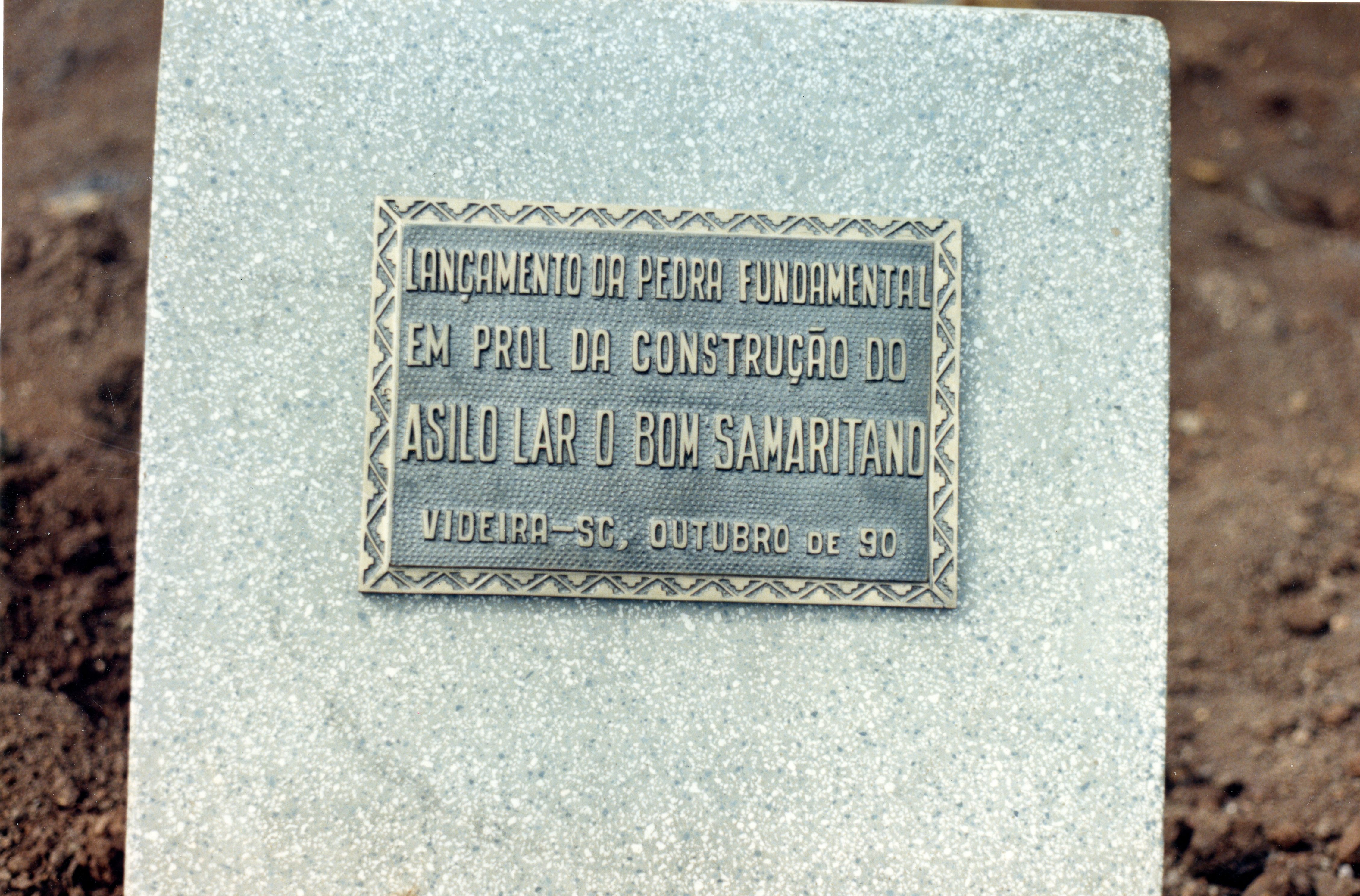 Uma placa comemorativa de metal fixada em um bloco de granito, marcando o lançamento da pedra fundamental do Asilo Lar O Bom Samaritano em Videira, Santa Catarina. A inscrição indica que o evento ocorreu em outubro de 1990, representando um marco importante para a infraestrutura social da cidade.