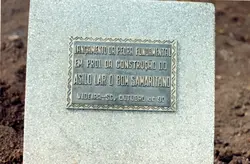 Uma placa comemorativa de metal fixada em um bloco de granito, marcando o lançamento da pedra fundamental do Asilo Lar O Bom Samaritano em Videira, Santa Catarina. A inscrição indica que o evento ocorreu em outubro de 1990, representando um marco importante para a infraestrutura social da cidade.