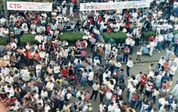 Uma vista aérea de uma grande multidão reunida em um evento público ao ar livre, possivelmente uma festividade cultural ou comunitária. Faixas suspensas mencionam 'CTG Pioneiros do Vale' e 'Tarde Nativista', sugerindo um evento tradicionalista gaúcho, provavelmente ocorrido no final do século XX.