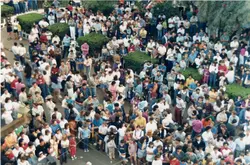 Uma vista aérea de uma grande multidão reunida em uma praça pública, possivelmente durante um evento comemorativo ou festividade local. As vestimentas e o estilo das pessoas sugerem um contexto brasileiro entre o final da década de 1980 e início da década de 1990.