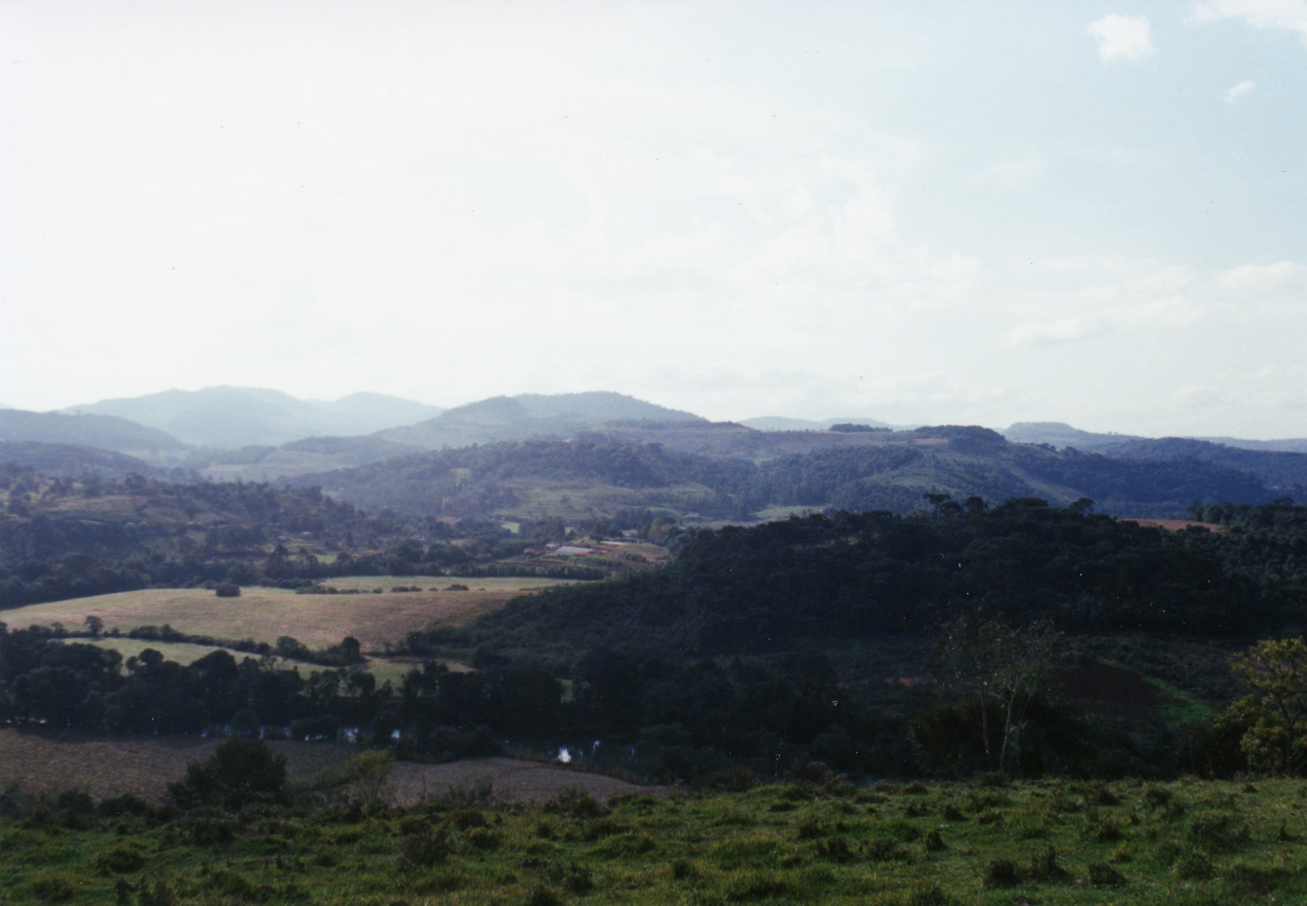 Uma paisagem rural brasileira com colinas suaves, campos de pastagem e áreas de mata nativa, possivelmente na região de Videira ou interior de Santa Catarina. A imagem apresenta uma topografia típica de relevo acidentado, com vegetação densa e áreas de cultivo, sugerindo um cenário de ocupação agrícola tradicional.