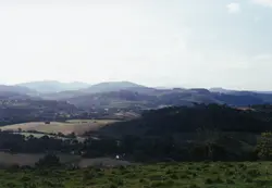Uma paisagem rural brasileira com colinas suaves, campos de pastagem e áreas de mata nativa, possivelmente na região de Videira ou interior de Santa Catarina. A imagem apresenta uma topografia típica de relevo acidentado, com vegetação densa e áreas de cultivo, sugerindo um cenário de ocupação agrícola tradicional.