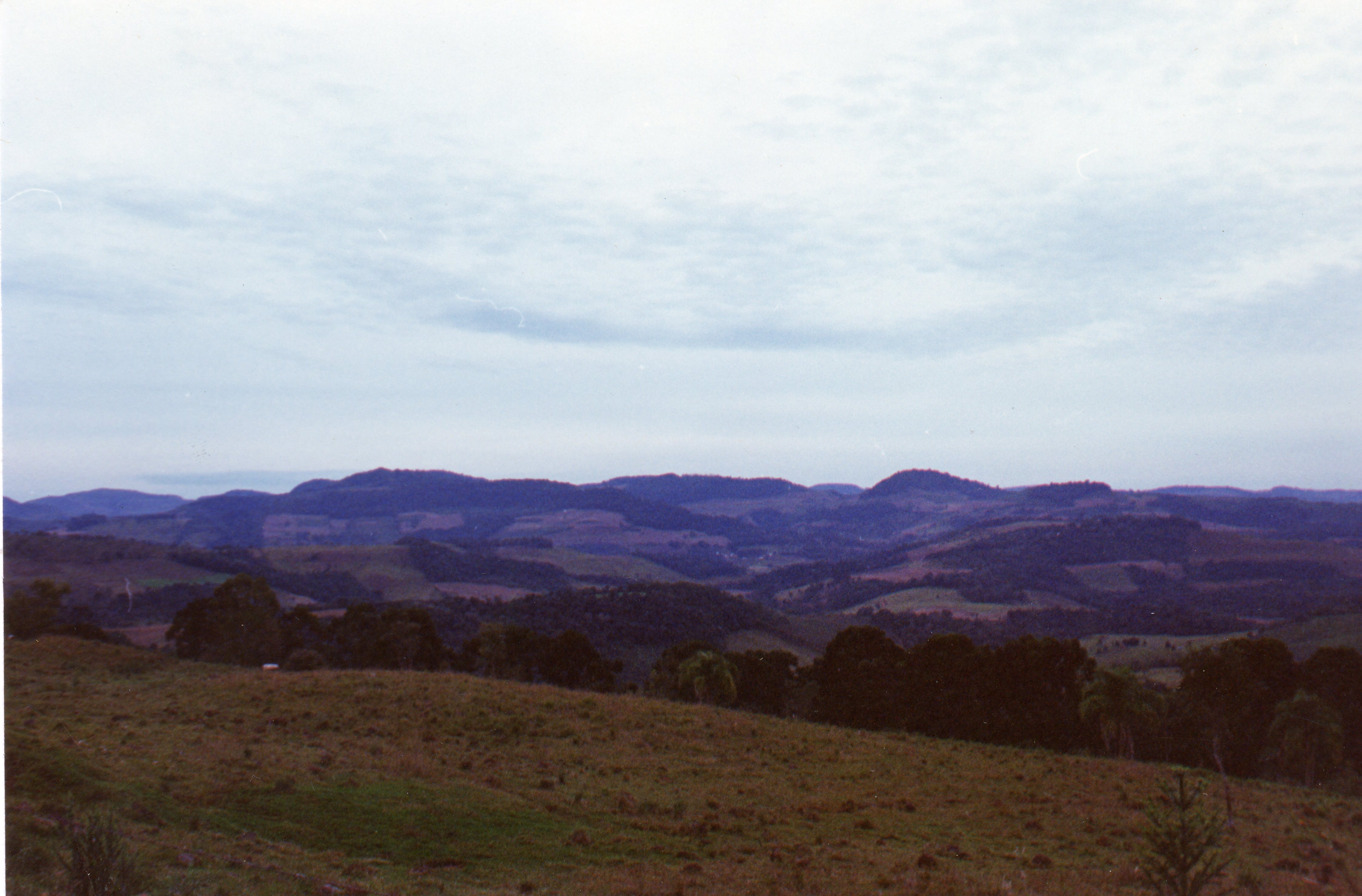 Uma paisagem rural brasileira com colinas onduladas e vegetação típica, possivelmente na região sul do país. A imagem apresenta um horizonte vasto sob um céu nublado, sugerindo uma área de relevo acidentado com campos e áreas de mata preservada.