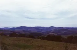 Uma paisagem rural brasileira com colinas onduladas e vegetação típica, possivelmente na região sul do país. A imagem apresenta um horizonte vasto sob um céu nublado, sugerindo uma área de relevo acidentado com campos e áreas de mata preservada.
