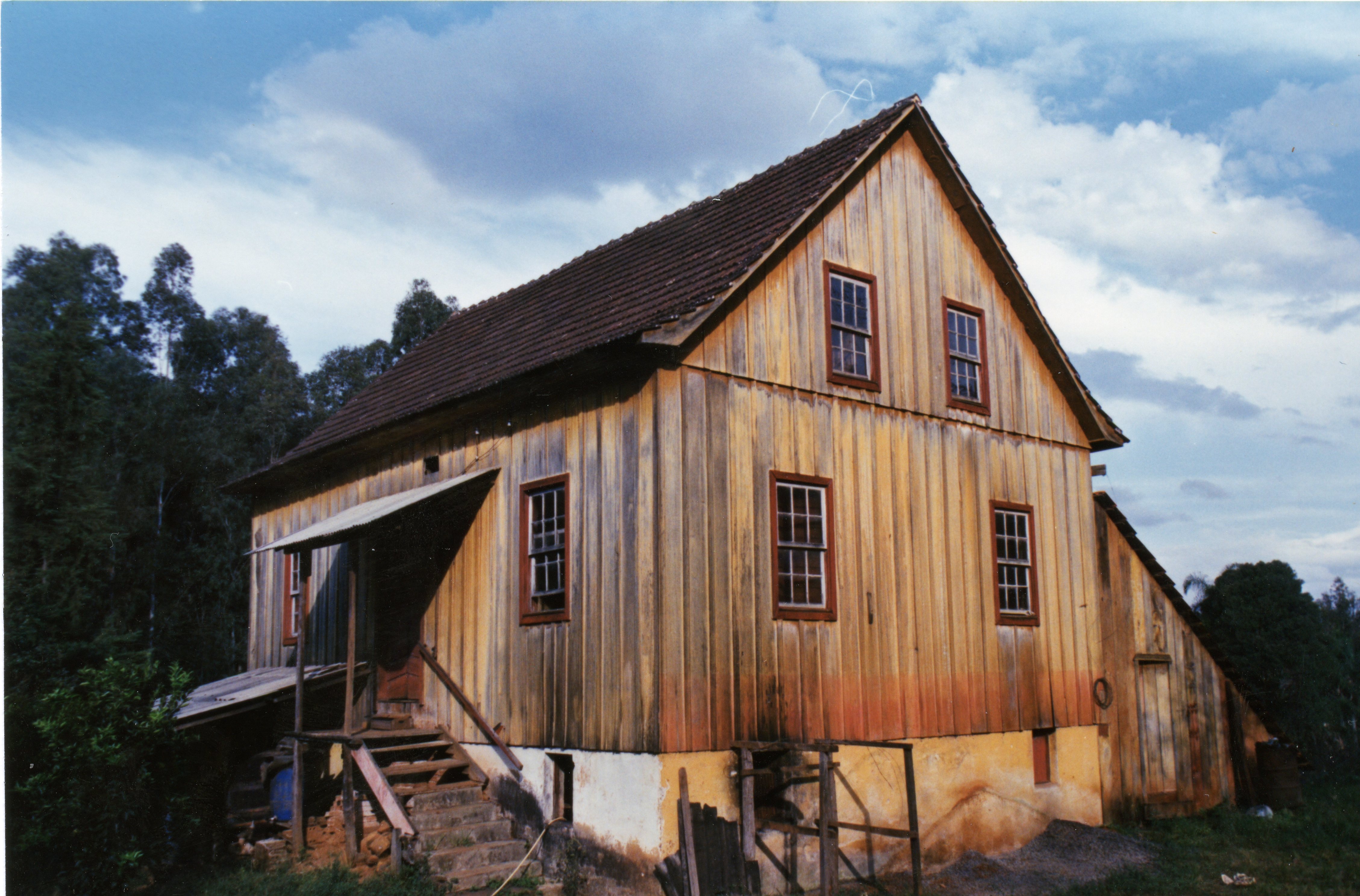 Uma casa de madeira tradicional com base de alvenaria, típica da arquitetura de colonização europeia no sul do Brasil. A estrutura apresenta telhado de duas águas e escadaria de acesso lateral, sugerindo uma construção rural antiga, possivelmente de meados do século XX.