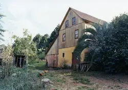 Uma construção rural de madeira e alvenaria, típica da arquitetura de colonização europeia no sul do Brasil, possivelmente um moinho ou celeiro antigo. O cenário inclui vegetação nativa, como araucárias ao fundo, e galinhas no terreno, sugerindo uma propriedade agrícola em uma área como Videira.