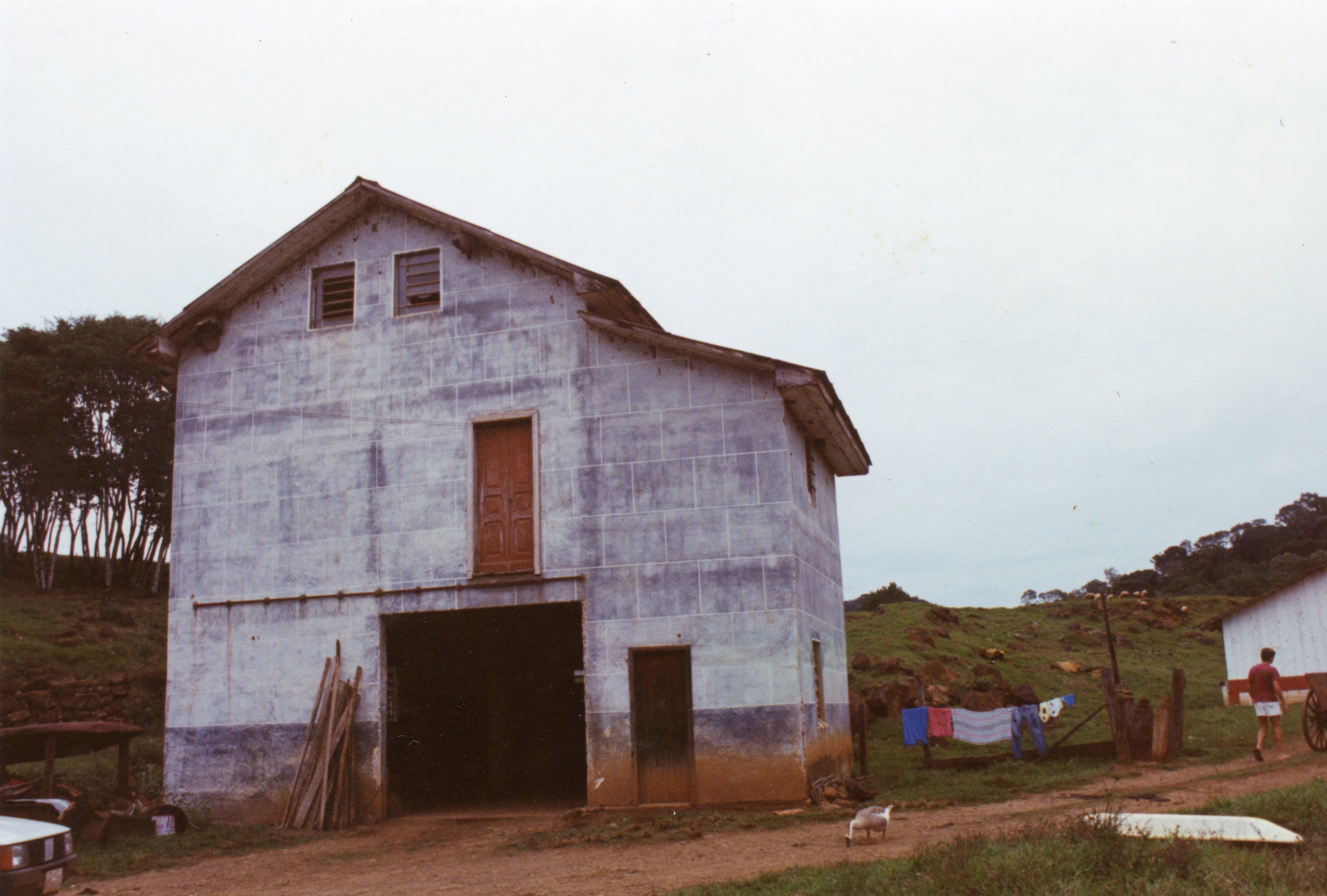 Uma construção rural de dois andares com paredes de alvenaria pintadas de azul claro, típica de propriedades agrícolas no interior do Brasil. A cena mostra um ambiente de fazenda com roupas estendidas em um varal, uma pessoa caminhando ao fundo e vegetação ao redor, sugerindo um estilo de vida simples e tradicional.