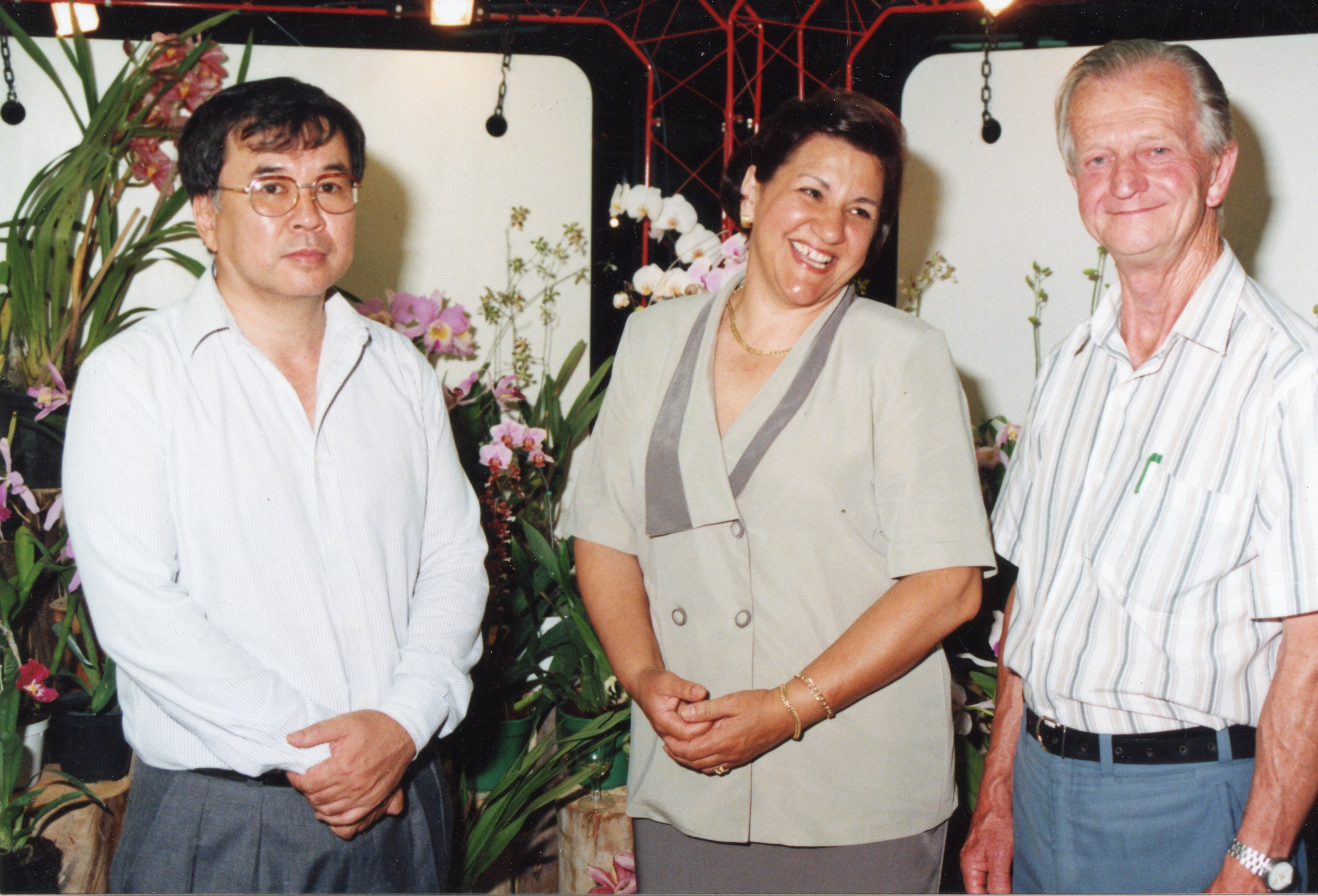 Uma fotografia colorida retratando três pessoas posando juntas em um ambiente interno decorado com orquídeas. O estilo das vestimentas e a qualidade da imagem sugerem um evento social ou exposição ocorrido provavelmente na década de 1990.