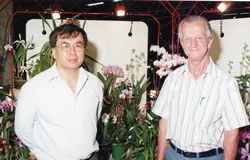Uma fotografia colorida retratando dois homens posando lado a lado em uma exposição de orquídeas. O ambiente sugere um evento social ou feira de flores, com iluminação artificial e estruturas metálicas ao fundo, possivelmente datando do final do século XX.