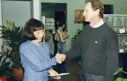 Uma fotografia colorida que mostra um aperto de mão entre um homem e uma mulher em um ambiente interno, possivelmente uma exposição ou evento de criadores de pássaros, dado o fundo com gaiolas. O estilo das roupas, como o suéter de lã e o corte de cabelo, sugere uma cena típica da década de 1990.