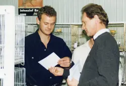 A imagem mostra dois homens examinando documentos ou papéis em um ambiente interno, possivelmente uma exposição ou competição de pássaros, dado o contexto das gaiolas ao fundo. A cena sugere um evento de criadores, com vestimentas casuais típicas das décadas de 1990 ou início de 2000.