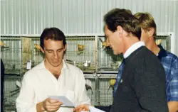Uma fotografia que mostra três homens em um ambiente interno, possivelmente uma exposição ou competição de canários, dado o grande número de gaiolas metálicas ao fundo. Os homens parecem estar analisando documentos ou fichas de avaliação, sugerindo um contexto de julgamento ou organização de evento ornitológico, provavelmente datado entre as décadas de 1980 e 1990.