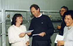 Uma fotografia colorida que retrata um momento de descontração entre três pessoas em um ambiente interno, possivelmente uma exposição ou clube de criadores de pássaros, dado o fundo com gaiolas. Os indivíduos parecem estar segurando papéis ou cartões, sugerindo uma premiação ou evento social, com vestimentas que remetem ao final da década de 1980 ou início da década de 1990.