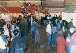 Uma cena interna movimentada, possivelmente uma feira de negócios ou exposição comercial, com pessoas circulando entre estandes. O ambiente apresenta balões decorativos e cartazes, sugerindo um evento público ou feira regional ocorrida provavelmente na década de 1990.