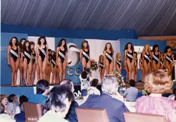 Uma fotografia de um concurso de beleza, provavelmente um Miss Santa Catarina, realizado em um ambiente coberto com decoração temática. As candidatas usam maiôs pretos com faixas identificando seus municípios, enquanto um público assiste e um cinegrafista da imprensa registra o evento.